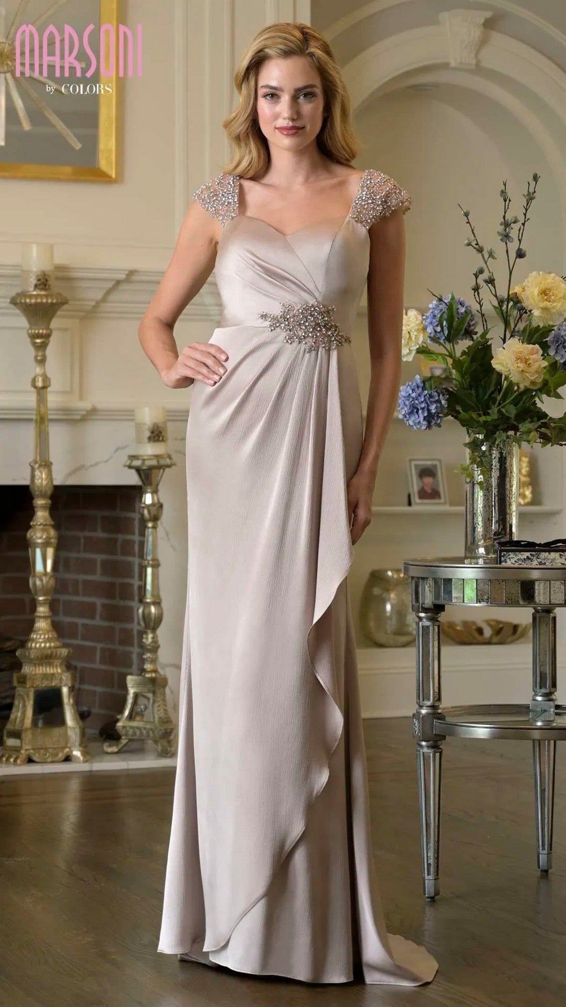 A woman poses gracefully indoors in a sand-colored Marsoni MV1377 long formal dress with beaded cap sleeves and a sparkling empire waist, standing by a fireplace and floral arrangement.